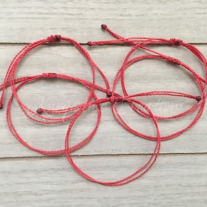 May include: Five red string bracelets with dark red beads at the ends. The bracelets are tied in a knot and are lying on a light wood surface.