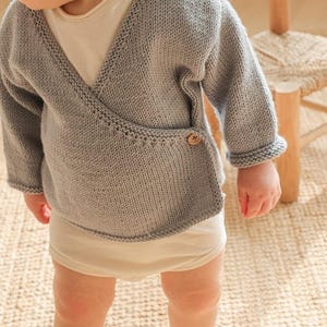 May include: A light gray knitted wrap sweater with long sleeves and a wooden button closure. The baby sweater has a V-neck design and ribbed cuffs. The baby is wearing a white onesie.