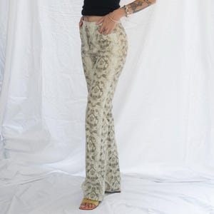 May include: A black sleeveless tank top paired with high-waisted trousers featuring a beige and dark grey snakeskin print pattern. The trousers have a straight-leg cut, front pockets, and a button closure at the waist, worn with yellow open-toe sandals.