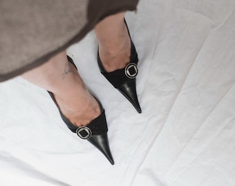 Zapatos de tacón destalonados de piel negra estilo Y2K con punta afilada y detalle de hebilla plateada.