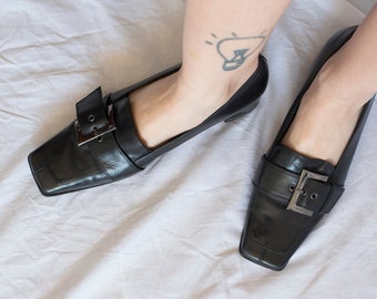 Puede incluir: Un par de mocasines de cuero negro con puntera cuadrada y hebilla plateada. Los mocasines tienen un tacón bajo.