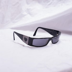 May include: Black sunglasses with rectangular lenses. The frames are black with a silver accent featuring a heart-shaped detail on the temples. The sunglasses are set against a white background.