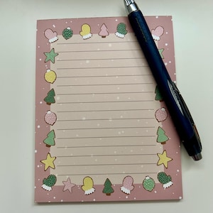 May include: Pink lined notepad with a Christmas themed border. The border features illustrations of mittens, baubles, Christmas trees, and stars. A blue pen is lying on top of the notepad.