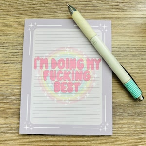 May include: A lined lavender notepad with the text "I'M DOING MY FUCKING BEST" in pink bubble letters, set against a rainbow background. A white and teal pen rests on the notepad, which has a decorative border.
