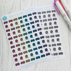 May include: Two sheets of stickers featuring hand-drawn yarn ball icons. The first sheet has a rainbow of colours, while the second sheet has a variety of shades of grey. The sheets are on a white wooden surface.