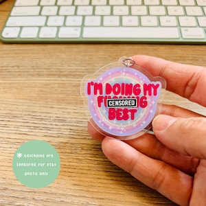 Peut inclure: Un porte-clés en acrylique transparent avec un fond arc-en-ciel et le texte "Je fais de mon mieux [CENSORED]" en lettres roses et blanches.