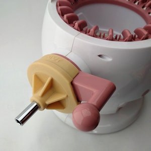 Könnte beinhalten: Nahaufnahme einer rosa und weißen Strickmaschine mit einer gelben und rosa Kurbel. Die Kurbel hat eine Metallstange, die aus ihr herausragt.