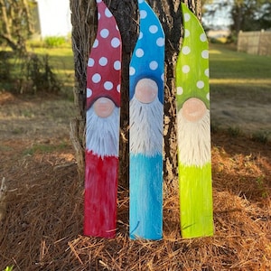 May include: Three colorful wooden garden gnomes leaning against a tree. The gnomes have pointed hats with white polka dots, painted faces, and white beards. The gnome bodies are red, blue, and green.