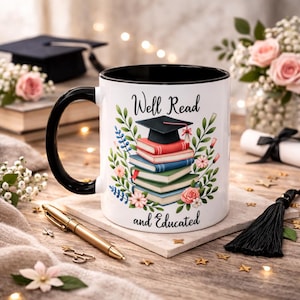 May include: White ceramic mug with a black handle and interior, inscribed with "Well Read and Educated." The design showcases a stack of books topped with a mortarboard, surrounded by floral motifs. A gold pen is placed nearby.