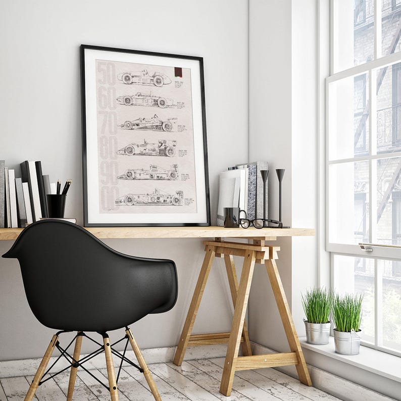 May include: Framed print featuring a series of race car designs from the 1950s to the 2000s. The black frame surrounds a beige background with the car illustrations and the years printed vertically on the left side. The print is displayed on a wooden desk.