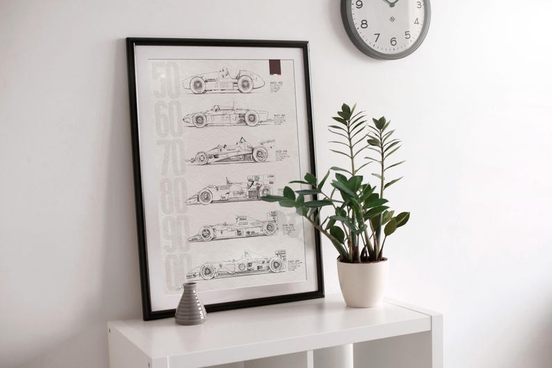 May include: Black framed print featuring detailed line drawings of vintage race cars from the 1950s to the 1990s. The print is displayed on a white shelf, next to a small gray vase and a potted plant. A clock hangs on the white wall.