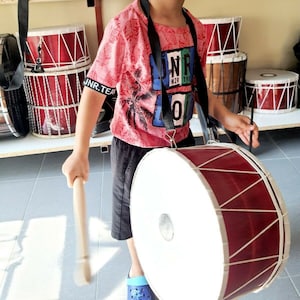 May include: A young person holding a large red and white drum with a wooden stick. The drum has a white head and a red body with white rope accents. The person is wearing a pink shirt with a palm tree design and the words "UNR N2R 19F" on the front.