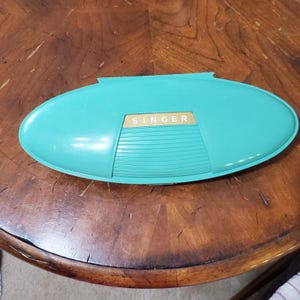 May include: An oval-shaped, teal-colored plastic storage case with a hinged lid. The center of the lid features a rectangular gold-colored panel with the word SINGER printed in white capital letters, above a section with horizontal ridges.