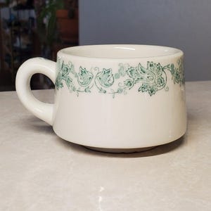 May include: A white ceramic mug with a black handle and the text "But First, Coffee" in a bold, black font. The mug is set against a blurred background, suggesting a cozy setting.