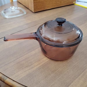 May include: A translucent purple glass saucepan with a matching lid featuring a circular textured pattern and a central knob handle. The saucepan has a long, flat, integrated glass handle extending from one side, providing a clear view of the vessel.