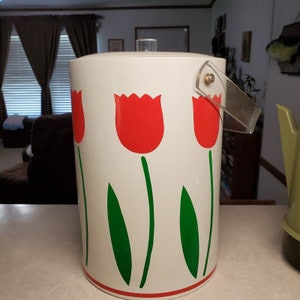 May include: A white ice bucket with a clear plastic handle and lid. The bucket is decorated with red tulip flowers with green stems and leaves. The bucket has a red band around the bottom.