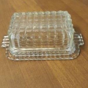 Puede incluir: Una mantequera de vidrio transparente con forma rectangular y un patrón texturizado en forma de rejilla. El plato tiene tapa y pequeñas asas a cada lado. El diseño es vintage y clásico.