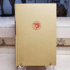 Puede incluir: Un libro antiguo con una cubierta beige y un lomo rojo. La portada presenta una ilustración roja de un pez dentro de un diseño circular. El libro se exhibe sobre un soporte transparente, posiblemente para la venta o exhibición.