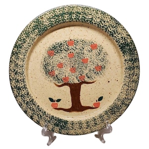 May include: A round ceramic plate with a hand-painted apple tree design. The tree has a brown trunk and branches with red apples. The rim is speckled green, and the background is beige.