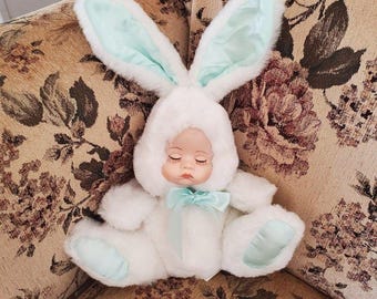 May include: A plush doll with a sleeping face wearing a white faux fur bunny suit. The suit features long ears with light mint green satin lining, a matching mint green satin bow at the neck, and mint green satin patches on the feet.