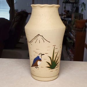 May include: A beige speckled ceramic vase with a scalloped rim. The exterior features a hand-painted design of a mountain range, a small bird with blue and brown plumage, and a green plant. The vase has a tapered neck and a wide base.
