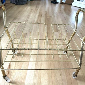 Może przedstawiać: Wózek na płyty winylowe w stylu vintage, wykonany z metalu w kolorze złotym, z prostokątnym drucianym stojakiem i przegrodami. Posiada zakrzywione uchwyty i toczy się na małych, przezroczystych kółkach.