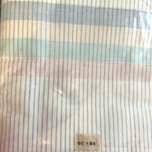 Pode incluir: Toalhas de mesa artesanais em embalagem plástica. Os lençóis têm listras verticais em azul claro, branco, verde e rosa. A etiqueta diz "60 x 84". A parte superior da embalagem diz "HANDCRAFTED TABLE LINENS".