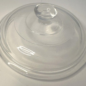 Puede incluir: Una tapa de vidrio transparente con un pequeño asa redondeada. La tapa es circular con anillos concéntricos y un área central texturizada. El diseño transparente permite la visibilidad del contenido. Un artículo de cocina.