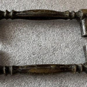 Puede incluir: Dos tiradores de armario antiguos de bronce. Cada tirador tiene un asa curvada con detalles decorativos y dos postes cilíndricos para el montaje. El acabado de bronce tiene un aspecto envejecido, lo que sugiere antigüedad. Ideal para restauración.