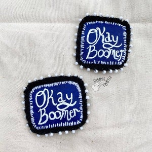 May include: Two blue and white embroidered patches with the text "Okay Boomer" on a white fabric background.