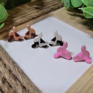 May include: A set of six cow-print stud earrings. Two are brown and white, two are black and white, and two are pink and white.