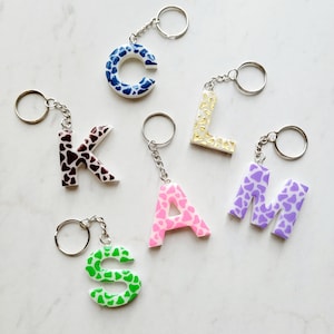 May include: Five keychains with different letters of the alphabet, each letter is a different color and has a cow print pattern. The letters are C, K, L, A, and M.