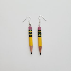 Puede incluir: Un par de pendientes de l&aacute;piz amarillo con detalles verdes y negros y gomas de borrar rosas. Los pendientes est&aacute;n sobre un fondo blanco.