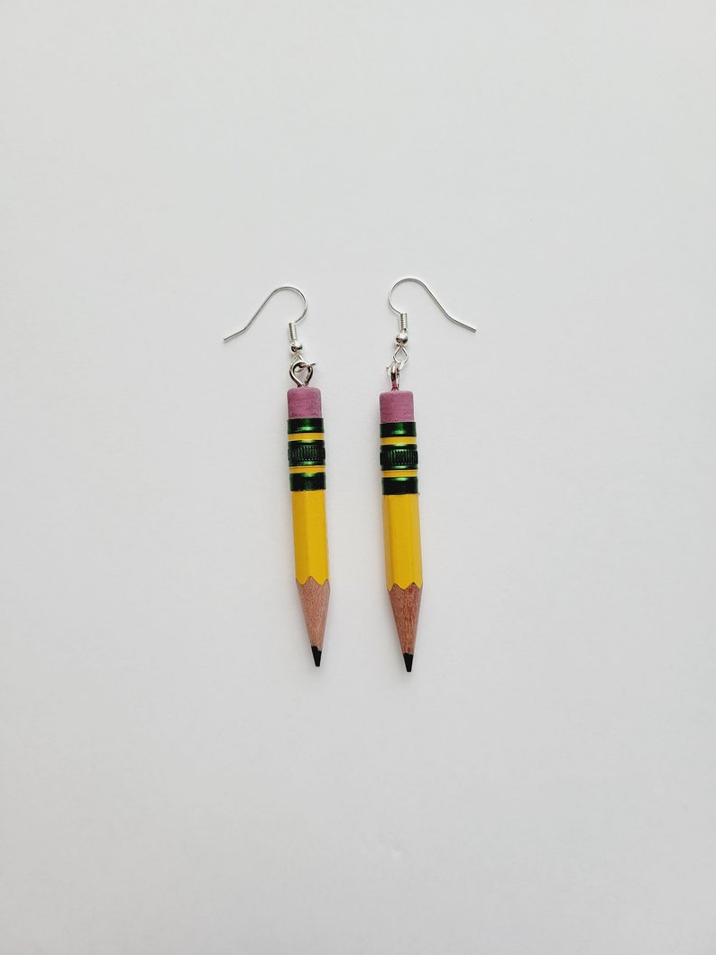 Puede incluir: Un par de pendientes de l&aacute;piz amarillo con detalles verdes y negros y gomas de borrar rosas. Los pendientes est&aacute;n sobre un fondo blanco.