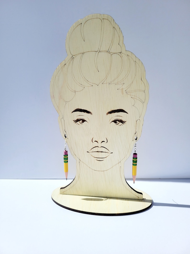 Puede incluir: Un expositor de pendientes de madera con una silueta de rostro femenino. El expositor sostiene un par de pendientes de l&aacute;piz amarillo con detalles verdes y rosas.
