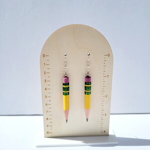 Puede incluir: Un par de pendientes de l&aacute;piz amarillo con detalles verdes y rosas. Los pendientes est&aacute;n colgados de un expositor de madera con una regla marcada en cent&iacute;metros.