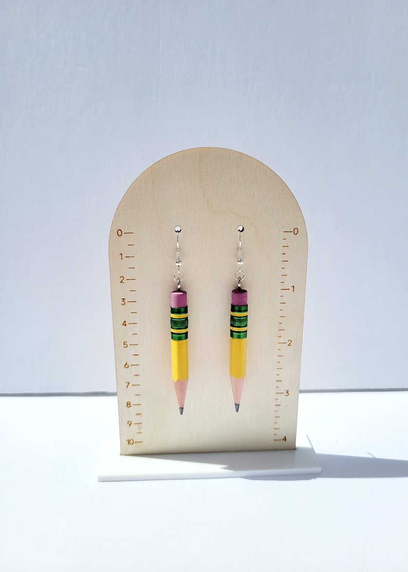 Puede incluir: Un par de pendientes de l&aacute;piz amarillo con detalles verdes y rosas. Los pendientes est&aacute;n colgados de un expositor de madera con una regla marcada en cent&iacute;metros.