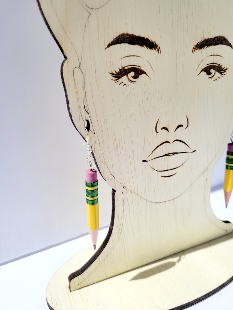 Puede incluir: Un par de pendientes de l&aacute;piz amarillo con detalles verdes y rosas. Los pendientes cuelgan de una cabeza de maniqu&iacute; de madera.