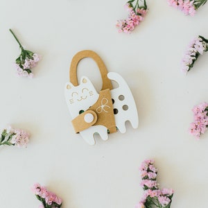 Pode incluir: Uma caixa de cartão branca em forma de gato com uma alça marrom e um padrão verde e branco dentro. A caixa é cercada por flores cor-de-rosa em um fundo branco.