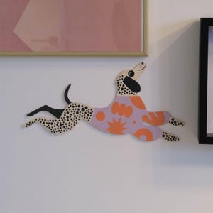 Puede incluir: Decoración de pared de madera con forma de perro corriendo. El perro tiene un cuerpo marrón claro con manchas negras, una cola negra y un atuendo con estampado lavanda y naranja. Está montado en una pared blanca.