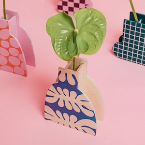 Peut inclure: Plusieurs vases décoratifs en bois de différentes couleurs et motifs, chacun contenant une feuille verte. L'un des vases présente un motif de feuilles bleu et blanc. Le fond est rose uni. Design moderne.