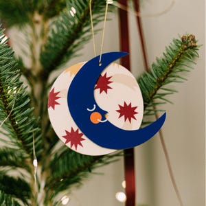 May include: A whimsical Christmas tree ornament featuring a smiling crescent moon in navy blue, with an orange nose and white eyes. The moon is layered over a cream-colored sphere decorated with red starbursts. A gold string is attached for hanging.