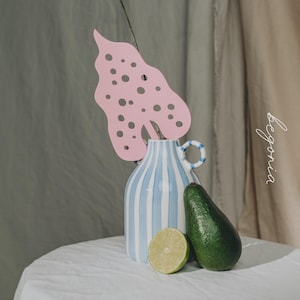 May include: A still life arrangement with a blue and white striped vase, a pink leaf cutout, a green avocado, and a lime half. The word "begonia" is written in cursive. The objects are on a white surface.