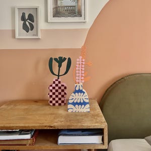 May include: Two decorative vases sit on a wooden nightstand. One vase is pink with a checkered pattern, and the other is blue with a white floral design. A green and pink abstract floral design sits atop each vase. The background features framed art and a peach-colored wall.
