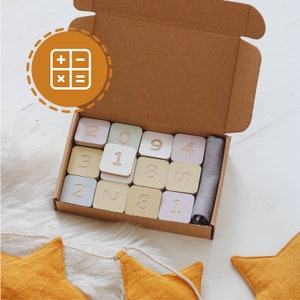 Könnte beinhalten: Eine Holzkiste gefüllt mit bunten Blöcken, auf denen jeweils eine Zahl aufgedruckt ist. Die Kiste ist geöffnet und die Blöcke sind sichtbar. Die Kiste ist aus Holz und hat eine hellbraune Farbe. Die Blöcke sind aus Holz und haben verschiedene Farben, darunter Rosa, Blau, Grün und Gelb. Die Zahlen auf den Blöcken sind weiß und gut lesbar.