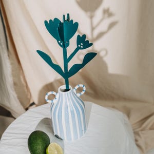 May include: A teal metal flower sculpture in a striped blue and white vase with two handles. The vase sits on a white surface with a lime and avocado. The flower has a detailed design with multiple petals and leaves.