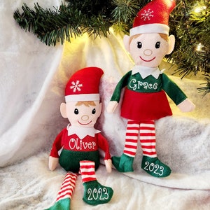 May include: Two personalized Christmas elves with red and green outfits, white striped stockings, and green shoes. The elves are sitting on a white fluffy surface. The elf on the left has the name "Oliver" embroidered on its shirt and the year "2023" on its shoe. The elf on the right has the name "Grace" embroidered on its shirt and the year "2023" on its shoe.