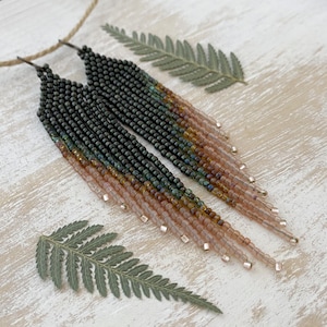 Può includere: Un paio di orecchini lunghi con perline a gradiente di colore verde, oro e rosa. Gli orecchini sono appesi a un filo e sono lunghi circa 10 cm.