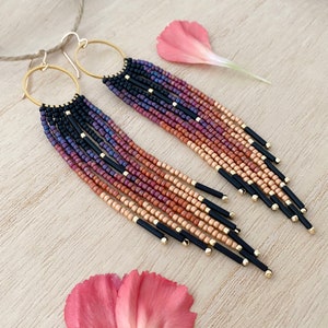 May include: A pair of long, beaded earrings with a gradient of colors from black to purple to orange to beige. The earrings are made with small seed beads and have gold hoops at the top.