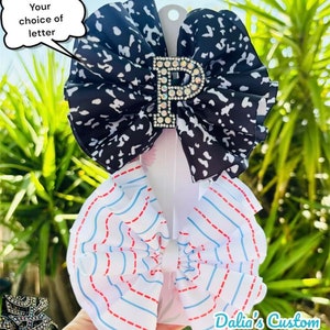 May include: Two hair bows, one with a black and white cow print and the other with red and blue stripes on a white background. The cow print bow has a rhinestone letter "P" in the center. Dalia's Custom Boutique is printed on the bottom right corner of the image.